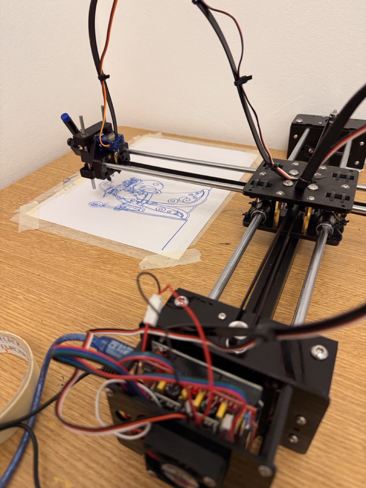 The first low-cost plotter drawing a character with a blue pen, wires and controller board visible.