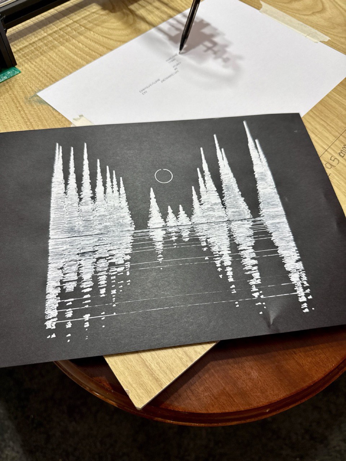 White gel pen on black paper: a landscape of trees reflected in water with a moon, Joy Division style.